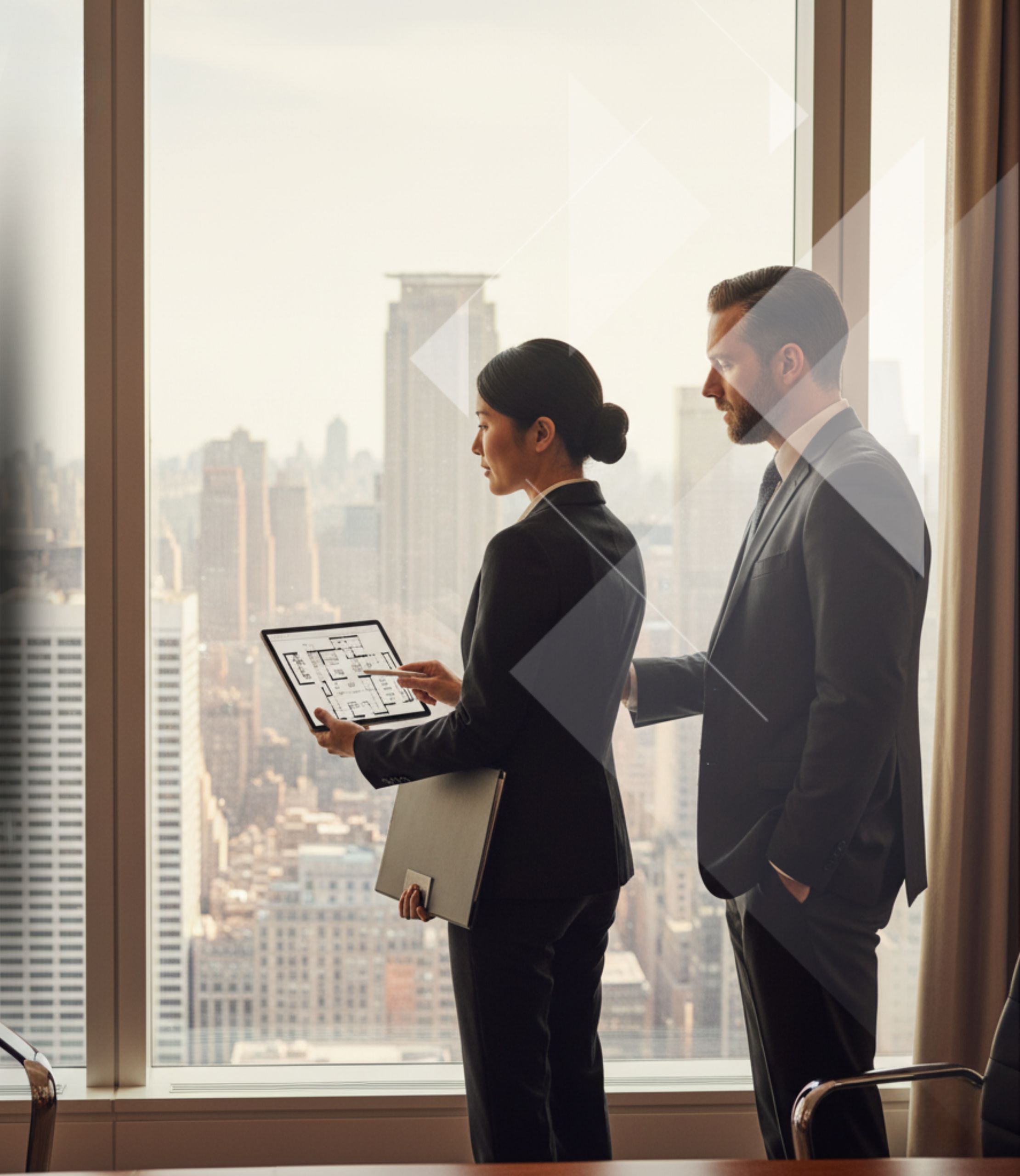 Two business professionals in suits stand by a large window in a high-rise office, looking out at a city skyline. The woman holds a tablet displaying architectural blueprints and points at it while the man looks on attentively.