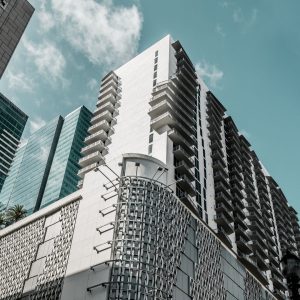 Looking up from the street at a modern white residential skyscraper featuring rows of balconies and a textured metallic lower facade, with glass office towers in the background.
