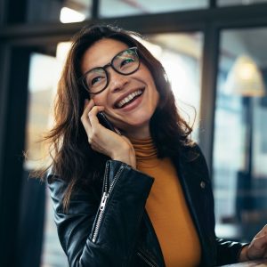 A smiling woman with dark hair and black-framed glasses wearing a yellow turtleneck and leather jacket while talking on her smartphone.
