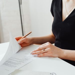 Close-up of a woman wearing a gold ring holding a wooden pencil over a paper contract at a white table.